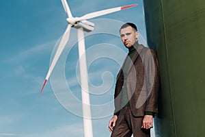 A man in a business suit with a green Golf shirt stands next to a windmill against the background of the field and the blue sky.