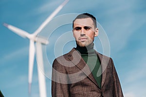 A man in a business suit with a green Golf shirt stands next to a windmill against the background of the field and the blue sky.