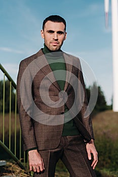 A man in a business suit with a green Golf shirt stands next to a windmill against the background of the field and the blue sky.