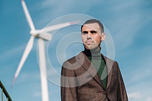 A man in a business suit with a green Golf shirt stands next to a windmill against the background of the field and the blue sky.