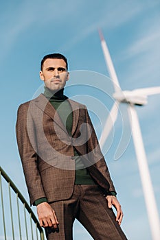 A man in a business suit with a green Golf shirt stands next to a windmill against the background of the field and the blue sky.