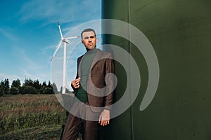 A man in a business suit with a green Golf shirt stands next to a windmill against the background of the field and the blue sky.