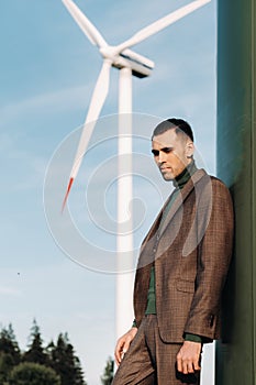 A man in a business suit with a green Golf shirt stands next to a windmill against the background of the field and the blue sky.