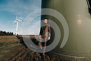 A man in a business suit with a green Golf shirt stands next to a windmill against the background of the field and the blue sky.