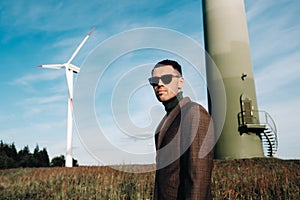 A man in a business suit with a green Golf shirt stands next to a windmill against the background of the field and the blue sky.