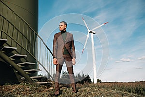 A man in a business suit with a green Golf shirt stands next to a windmill against the background of the field and the blue sky.