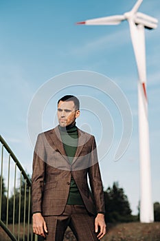 A man in a business suit with a green Golf shirt stands next to a windmill against the background of the field and the blue sky.