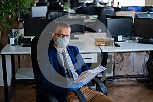 A man in a business suit and medical mask reads a paper report in an empty open space office. Social distance and