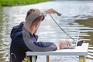 A man in a business suit sits at a white table in a swamp, in an outstretched left hand holds a telephone receiver