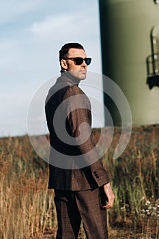 A man in a business suit stands next to a windmill against a field and blue sky.Businessman near the windmills.Modern concept of