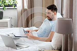 man with calculator, bills and laptop works in bed