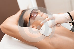 The man came to the procedure of laser hair removal. The doctor treats his armpit with a special apparatus.