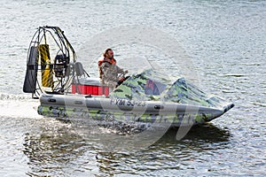 Man in camouflage uniforms and life jacket riding airboat along water surface. Inflatable rubber air boat sail on river