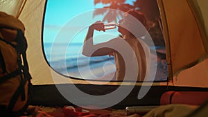 A man is on a camping vacation relaxing by the ocean. Young man sitting near a tent at sunset on the beach drinking