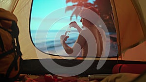 A man is on a camping vacation relaxing by the ocean. Young man sitting near a tent at sunset on the beach drinking