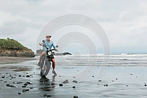 Man in cap riding motorcycle on beach. Moto cross dirtbiker on beach sunset on Bali. Young hipster male enjoying freedom