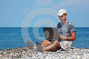 Man in cap sitting on beach with laptop on knees