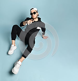 Man in cap, sunglasses, black tracksuit, white sneakers. He sitting on floor, gesticulating, posing on blue background. Close up