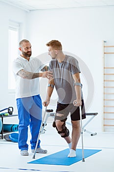 Man after car accident in an orthosis and on crutches learning to walk in the clinic, helpful therapist near him