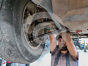 A man in a car dealership is repairing a broken car on a lift. A mechanic in overalls is repairing a car. Vehicle repair