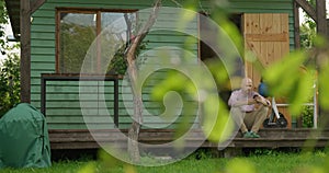 A man carpenter is sitting on the veranda of his tiny carpentry workshop resting