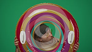 Man in casual clothing holds inflatable tube ring and looking through it with amazed face expression. Isolated on green