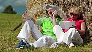 Man with cell phone and woman with Tablet PC sits near a haystack