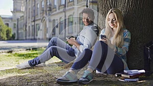 Man with cellphone sitting under tree and looking at girl using phone, affection