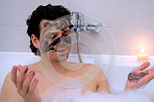 Man with charcoal face mask lying in bath with foam candle meditating at home.