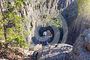 Man in a checkered blue shirt and a cap stands on the edge of a cliff of a deep canyon, looks down in surprise and a beautiful