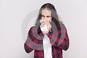 Man in checkered red shirt sneezes in white tissue, suffers from rhinitis and running nose.