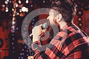 Man in checkered shirt holds microphone, singing song, karaoke club background. Guy likes to sing in dark karaoke hall