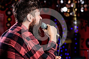 Man in checkered shirt holds microphone, singing song, karaoke club background. Guy likes to sing in dark karaoke hall