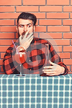 Man in checkered shirt near bottle, brick wall and blue tablecloth background. Barman with beard on shocked face holds