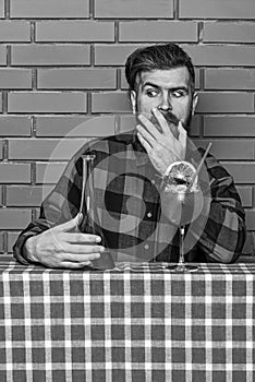 Man in checkered shirt near bottle, brick wall and blue tablecloth background. Barman with beard on shocked face holds