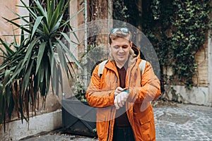 Man checking the time on his watch. Young stylish man wearing in travel orange jacket, standing on the street on city