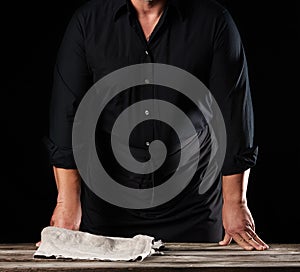 Man chef in a black shirt and apron stands near a wooden table, rested his hands