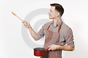 Man chef or waiter in striped brown apron, shirt pointing aside red empty stewpan wooden spoon on copy space isolated on