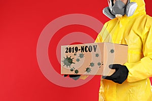 Man in chemical protective suit holding cardboard box on red background, closeup view with space for text. Coronavirus outbreak