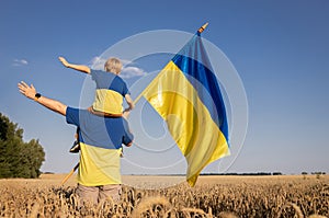 man and child, who sits on shoulders, stand with their backs proudly holding Ukrainian flag among wheat field