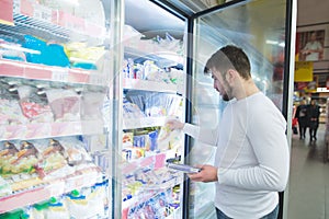 A man chooses frozen foods from shelves in a refrigerator in a supermarket. A man buys products in the store