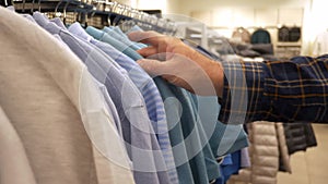 A man chooses a T-shirt in a store. A man's hand moves hangers with clothes.