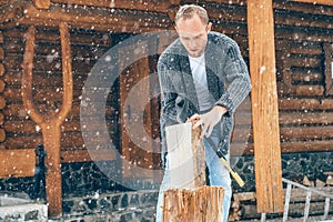 Man chopping wood on snowy yard for a house fireplace with heavy snowflakes background . Winter countryside holidays concept image