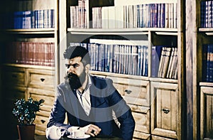 Man in classic suit sits in vintage interior, library, book shelves on background. Oldfashioned man near cup with tea