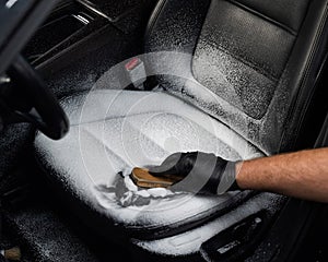 Man cleaning black leather car seat with brush and cleaning foam.