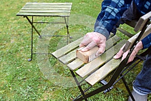 Man is cleaning and restoring wooden outdoor furniture, sanding the weathered wood to remove algae before oiling or painting for a