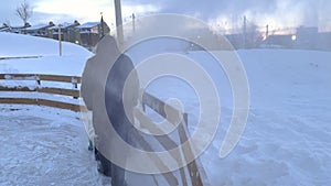 Man cleaning snow using snowblower near wooden fence on city street in winter.