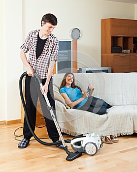 Man cleaning with vacuum cleaner while wife lying with eBook