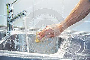 A man cleaning and washing the sink in the kitchen. One arm, one hand holds a sponge while soaping the whole surface. Water