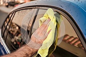 A man cleaning a window of a blue car with microfiber cloth at sunset. Manual car washing concept.